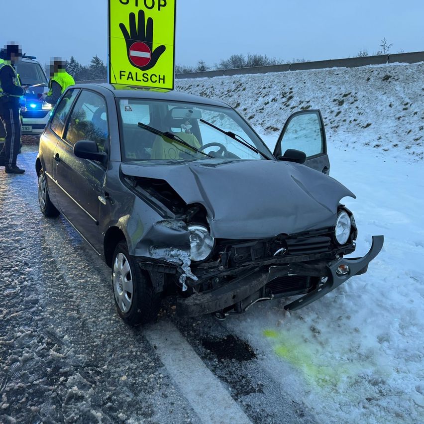 Ein schwarzes Auto mit beschädigtem vorderen Stoßfänger steht am Straßenrand. Darüber befindet sich ein STOP-Schild. Zwei Personen sind in der Nähe, eine trägt eine reflektierende Weste.
