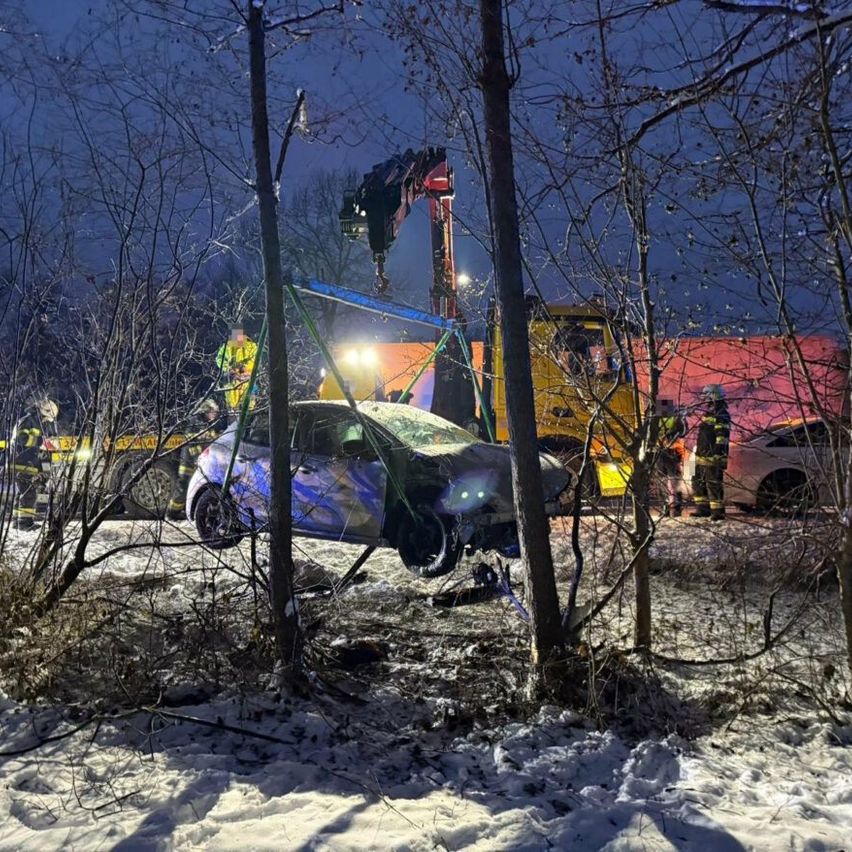 Ein beschädigtes blaues Auto wird von einem Kran in einem verschneiten Gebiet gehoben, mit anwesenden Rettungsfahrzeugen und Personal.