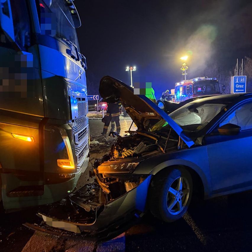 A severely damaged car with its hood open is at a collision with a truck at night. Firefighters are on the scene.