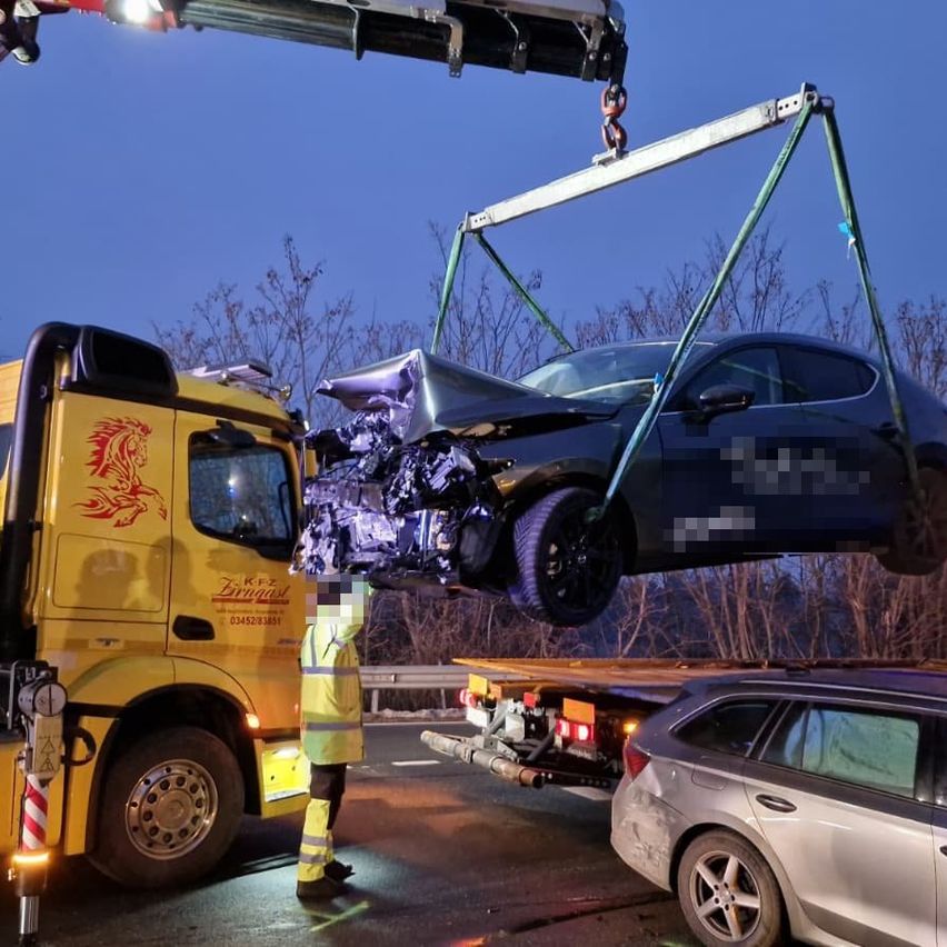 A wrecked black car is being lifted by a crane from a road. A yellow truck with a logo on the side is parked on the road. A person is standing nearby.