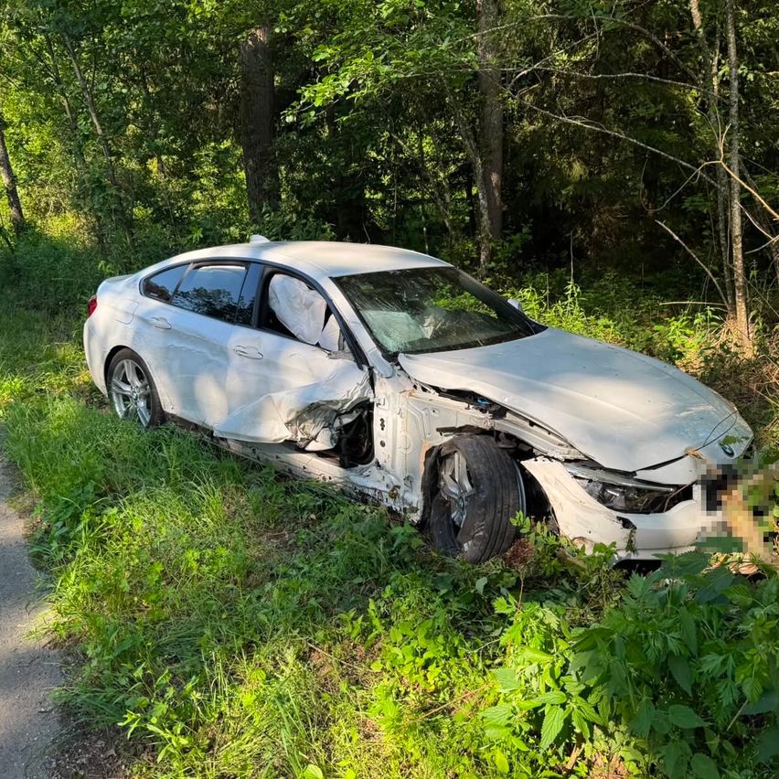 A white BMW is wrecked off the road, surrounded by greenery and trees. The front is heavily damaged, with a tire missing.