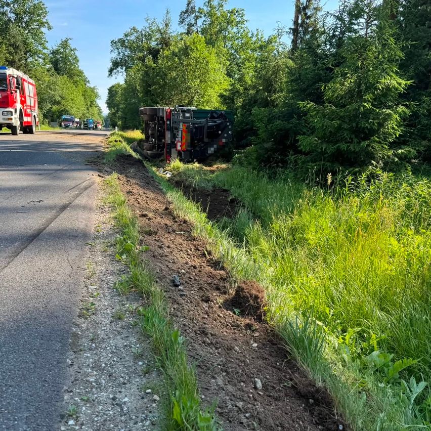 A red fire truck is parked by the roadside with a rolled-over vehicle on the grassy side. Trees surround the area, and a few cars are driving on the road.
