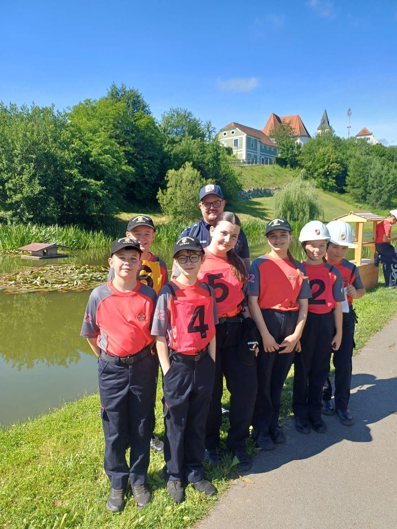 Eine Gruppe von Kindern mit nummerierten Shirts steht am Teich, mit einem Mann mit Brille und einem Haus im Hintergrund.