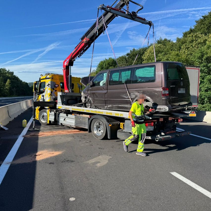 Ein Abschleppwagen hebt einen beschädigten Van von der Straße. Ein Mann in Sicherheitskleidung geht in der Nähe. Bäume und ein blauer Himmel mit Wolken sind im Hintergrund.