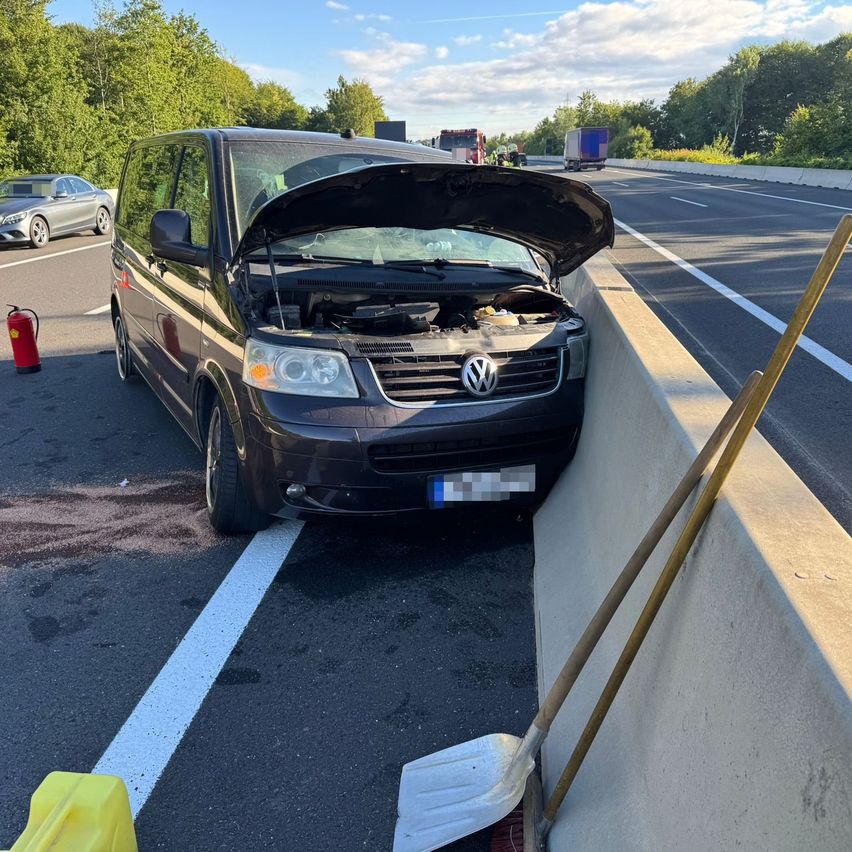 Ein schwarzer Volkswagen-Transporter mit offenem Motorhaube steht am Straßenrand neben einer Betonbarriere. Ein Feuerlöscher ist in der Nähe, und der Verkehr bewegt sich dahinter.