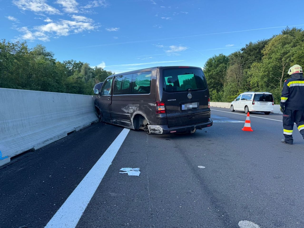 Ein brauner Volkswagen-Transporter ist auf der Straßenseite gegen eine Barriere gecrasht. Ein Verkehrskegel steht auf der Straße. Bäume und blauer Himmel sind im Hintergrund.