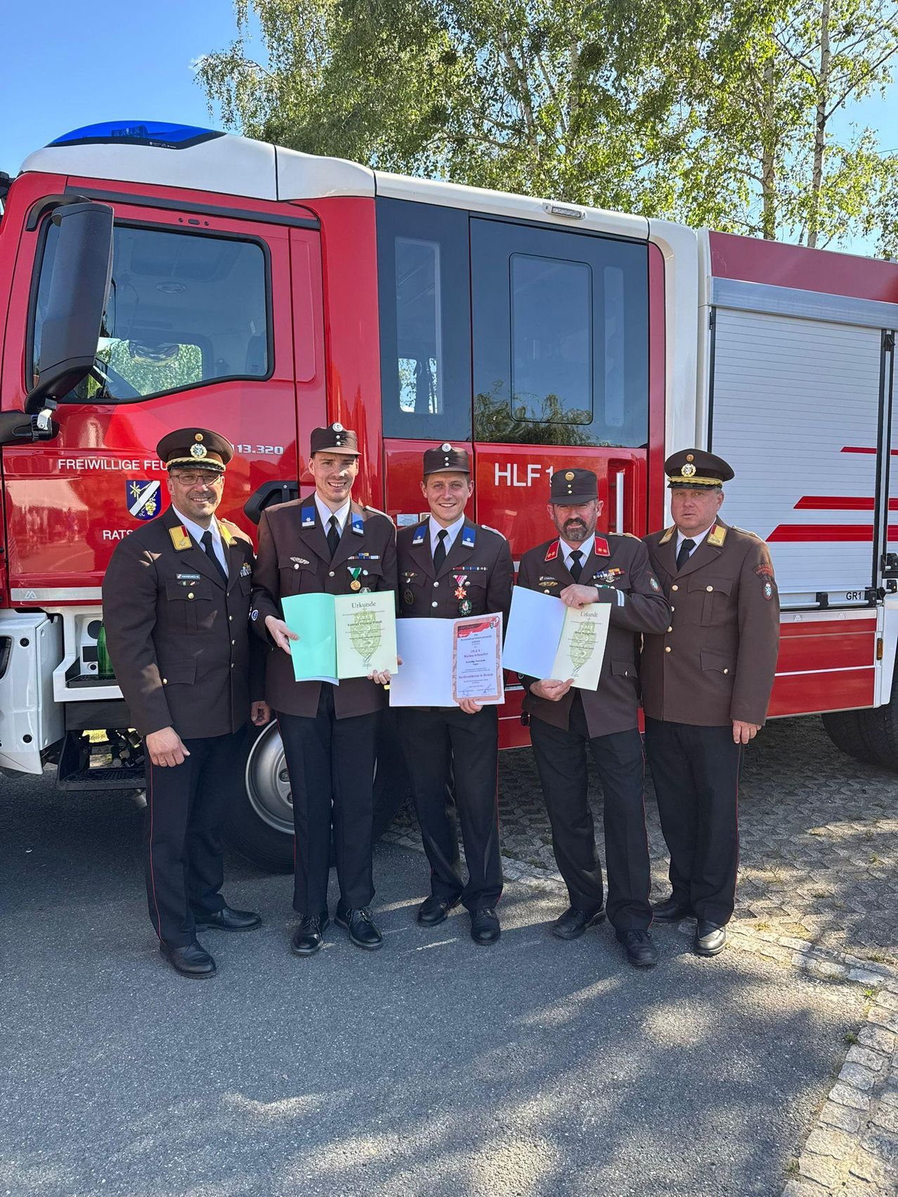 Fünf Männer in Uniform stehen vor einem roten Feuerwehrauto. Sie halten Papiere in der Hand und scheinen für ein Foto zu posieren.