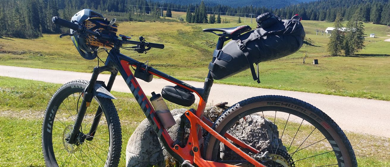 Ein Mountainbike mit orange-schwarzem Rahmen steht auf einem Grasfeld, mit einer Wasserflasche am Rahmen befestigt und einem großen schwarzen Rucksack hinten. Das Fahrrad befindet sich auf einem Felsen in der Nähe einer Straße, mit einem bewaldeten Berg im Hintergrund.