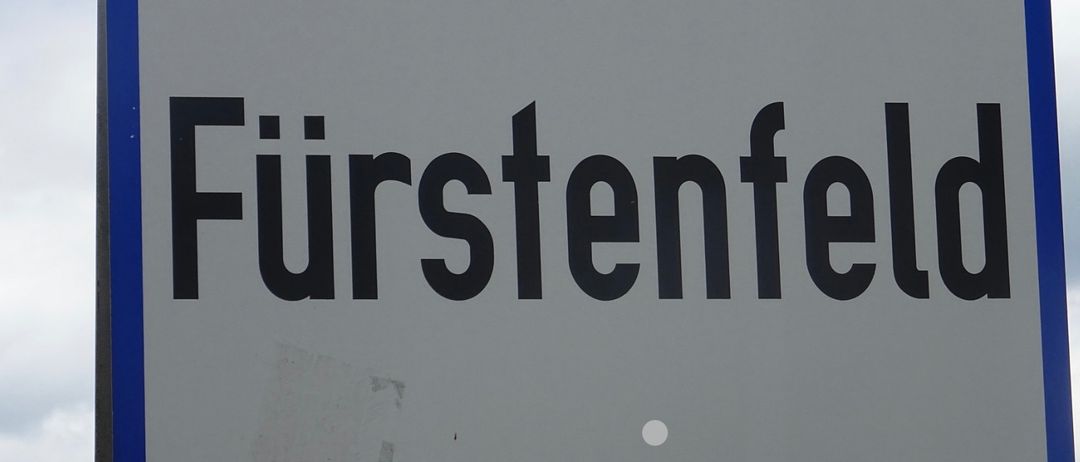 Ein Schild für den Ort Furstenfeld ist auf einem grauen Hintergrund mit blauer Einfassung angebracht. Der Text ist fetter schwarzer Druck.