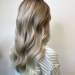 Eine Frau mit langen, hellblonden Haaren in lockeren Wellen steht vor einer weißen Wand. Sie trägt ein hellblau-weiß gestreiftes Hemd.