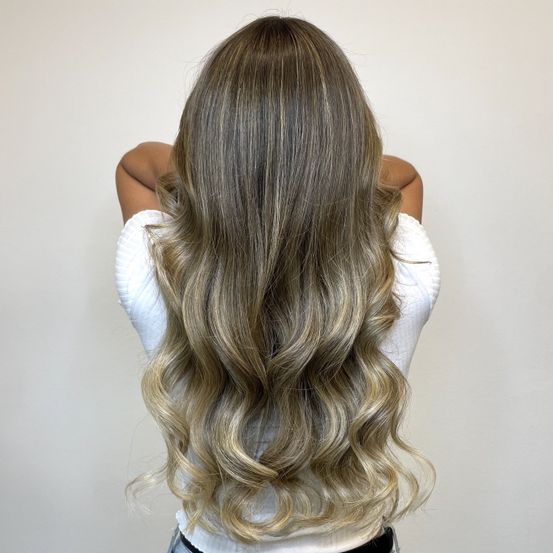 Eine Frau mit langen, welligen, blonden Haaren steht mit dem Rücken zur Seite und trägt ein weißes Oberteil. Der Hintergrund ist eine schlichte, helle Wand.