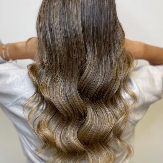 Eine Frau mit welligem, langem, blondem Haar, das eine graue Bluse trägt und ihre Haare mit den Händen hält.