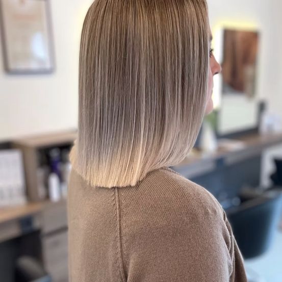 Eine Frau mit schulterlangen, blonden Haaren steht in einem Salon und schaut nach rechts. Hinter ihr ist ein Schreibtisch mit verschiedenen Gegenständen zu sehen. An der Wand hängen ein gerahmtes Bild und ein Spiegel.