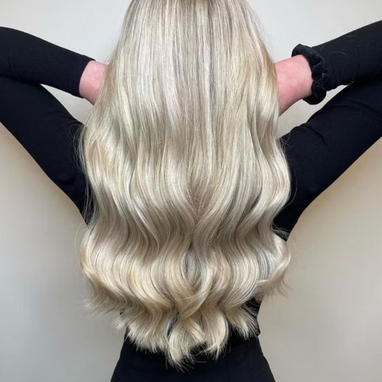 Eine Frau mit langen, glänzenden, blonden Haaren in lockeren Wellen wird von hinten gesehen, sie trägt ein schwarzes Langarm-Oberteil.