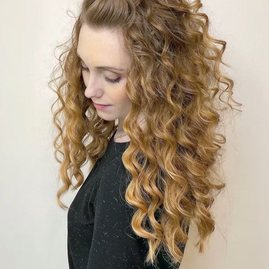 Eine Frau mit lockigem Haar, die nach unten schaut, trägt ein schwarzes Oberteil vor weißem Hintergrund.