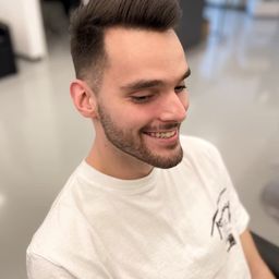 Ein Mann mit kurzem Haarschnitt und Bart lächelt in einem weißen T-Shirt in die Kamera. Der Hintergrund scheint ein professioneller Salon zu sein.