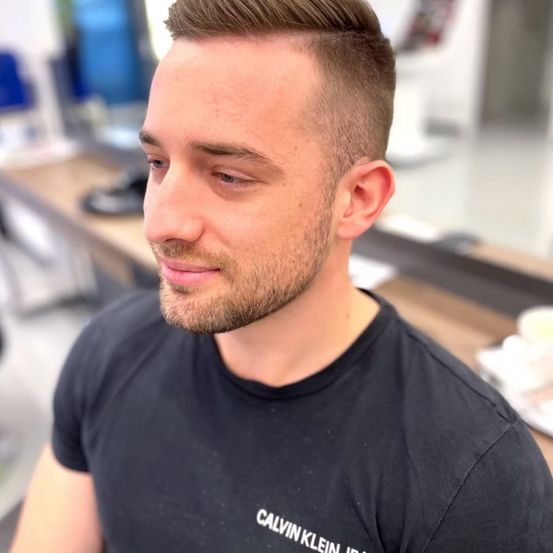 Ein Mann mit kurz geschnittenen Haaren und Bart, der ein schwarzes Calvin Klein T-Shirt trägt, lächelt in einem Salon.