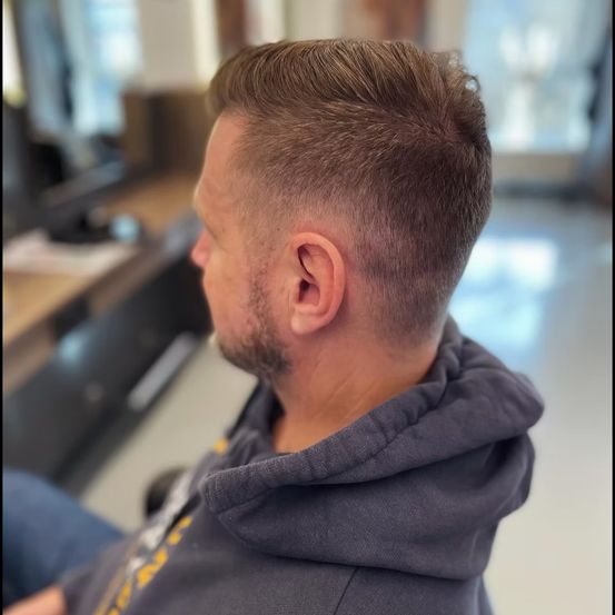 Ein Mann mit kurzen braunen Haaren und Bart sitzt in einem Raum und trägt eine graue Kapuzenjacke.