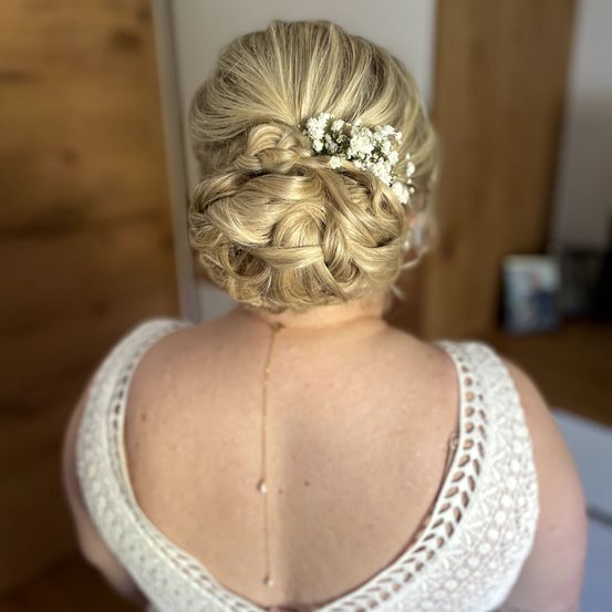Die rückseitige Ansicht einer Frau mit blonden Haaren in einem eleganten Dutt, geschmückt mit weißen Blumen und einer goldenen Halskette. Sie trägt ein weißes Kleid mit Rückendesign.