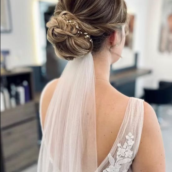 Eine Frau mit einer Brautfrisur und Schleier steht in einem Salon, der Rücken ist zur Seite gewandt. Der Schleier fließt über ihren Rücken.