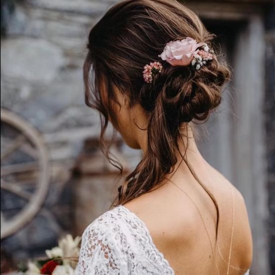 Eine Braut steht mit dem Rücken zugewandt, in einem weißen Spitzenkleid und mit Blumen im Haar. Der Hintergrund zeigt eine rustikale Umgebung mit einer Steinmauer und einem Rad.