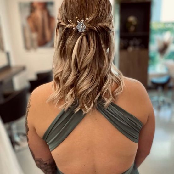 Eine Frau mit blonden Haaren in einem Salon. Sie trägt ein graues Kleid und hat eine blumige Haarspange. Im Hintergrund sind Stühle und ein Regal mit Gegenständen zu sehen.