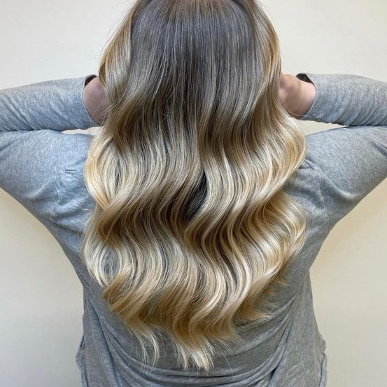 Eine Frau mit langen, welligen blonden Haaren in einem lockeren, natürlichen Look, trägt ein graues Langarmshirt.