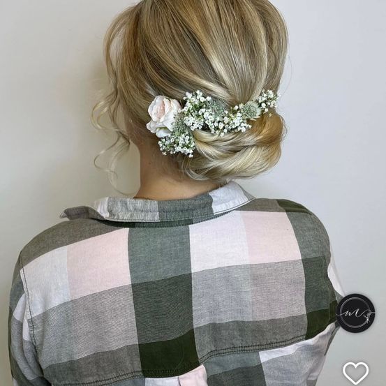 Eine Frau mit blondem Haar in einer Hochsteckfrisur, geschmückt mit einem rosa-weißen Blumenhaarzubehör. Sie trägt ein kariertes Hemd.