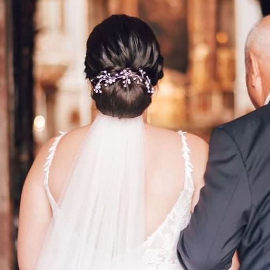 Eine Braut mit Dutt-Frisur und rosa Blumenklammer steht mit dem Rücken zur Seite, begleitet von einem älteren Mann im Anzug.