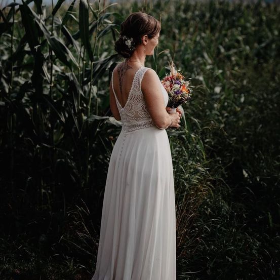 Eine Braut in einem weißen Kleid steht in einem Maisfeld und hält einen bunten Blumenstrauß. Der hohe Mais umgibt sie.