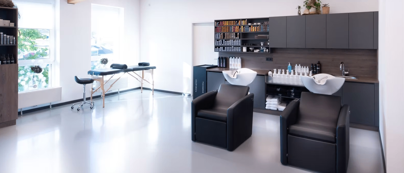 Ein Salon mit einem Massagetisch, zwei Stühlen und einem Waschbecken. Regale gefüllt mit Flaschen und Produkten. Der Raum hat Holzbalken, ein Fenster und einen weißen Boden.