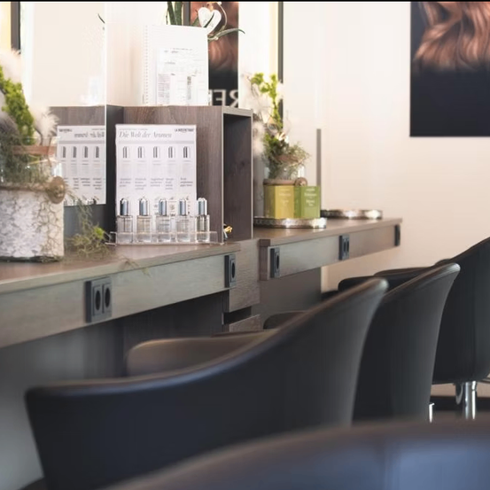 Ein modernes Salon-Interieur mit schwarzen Lederstühlen und einer Holztheke. Topfpflanzen und Produktdisplays schmücken die Theke. Ein gerahmtes Porträt hängt an der Wand.