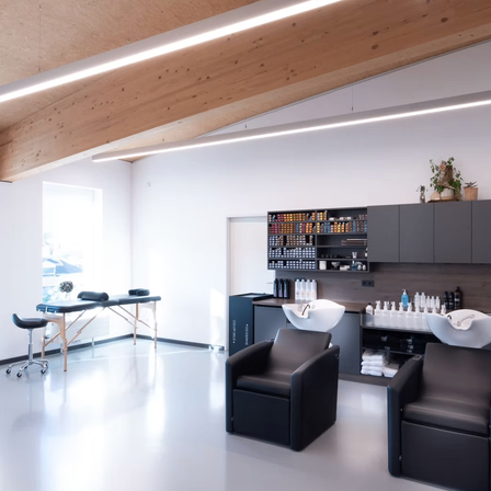 Ein moderner Salon mit weißen Wänden, einer Holzdecke und einem Schreibtisch mit Stuhl. Es gibt zwei schwarze Stühle und ein Waschbecken mit Flaschen auf der rechten Seite. Ein Regal mit verschiedenen Gegenständen ist an der Wand.