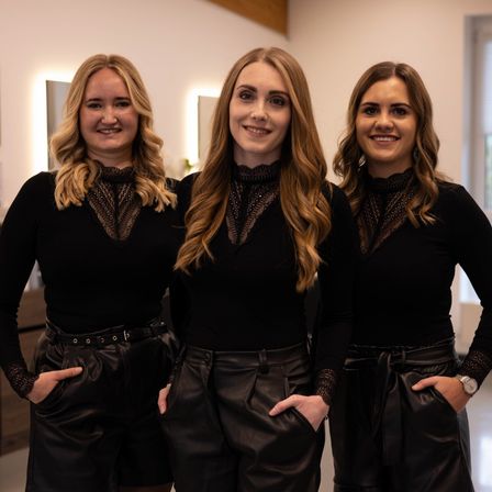 Drei Frauen in schwarzer Kleidung stehen zusammen und lächeln für die Kamera. Sie haben blonde Haare und tragen Lederhosen. Der Hintergrund ist hell, mit einer weißen Wand und einem Fenster.