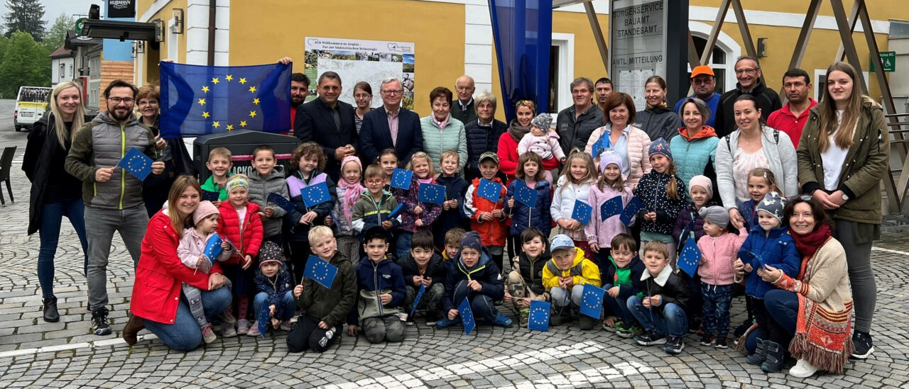 Eine Gruppe von Menschen, darunter Erwachsene und Kinder, posiert für ein Foto vor einem Gebäude mit einer europäischen Flagge. Einige halten kleine europäische Flaggen. Das Gebäude hat ein Wappen und eine blaue Flagge mit Sternen.