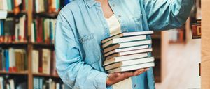 Eine Person in einem Jeanshemd hält einen Stapel Bücher in einer Bibliothek. Der Hintergrund zeigt Regale voller Bücher.