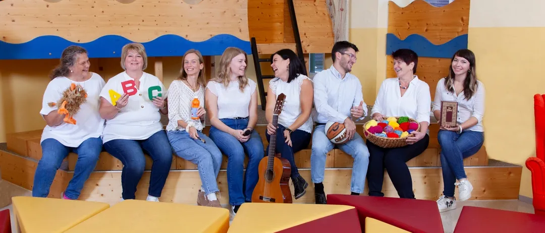 Eine Gruppe lächelnder Menschen sitzt auf Holzbänken, einige halten eine Gitarre, einen Basketball und einen Korb mit Wolle. Im Hintergrund befindet sich eine Holzkonstruktion mit einer Leiter und Fenstern.