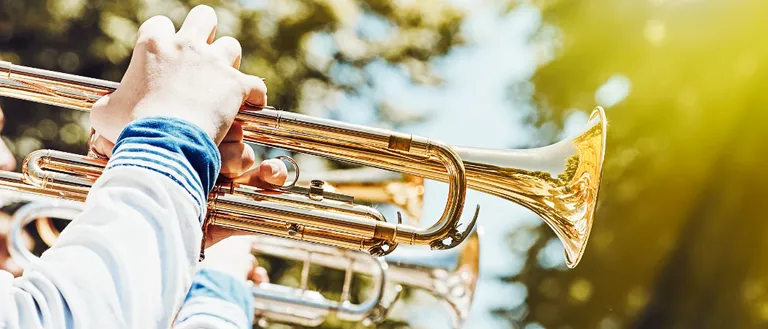 Die Hände eines Musikers liegen auf einer Messingtrompete, bereit zum Spielen. Die Trompete erstrahlt in goldenem Glanz, und ein verschwommener Hintergrund zeigt Bäume.
