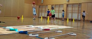 Indoor-Sporthalle mit Kindern, die Basketball spielen, ein Junge springt zum Werfen. Verschiedene farbige Bälle und Hütchen verstreut auf dem Boden.