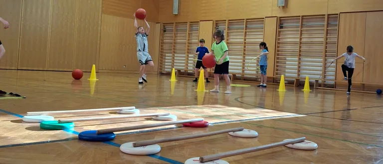 Indoor-Sporthalle mit Kindern, die Basketball spielen, ein Junge springt zum Werfen. Verschiedene farbige Bälle und Hütchen verstreut auf dem Boden.