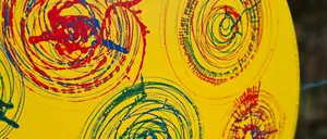 Ein gelbes rundes Objekt mit mehreren Spiralen in Rot, Blau und Grün bemalt. Die Spiralen sind unregelmäßig und haben unterschiedliche Dicke. Der Hintergrund ist gelb.