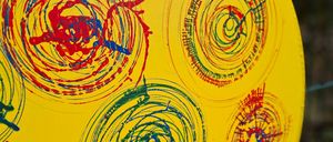 Ein gelbes rundes Objekt mit mehreren Spiralen in Rot, Blau und Grün bemalt. Die Spiralen sind unregelmäßig und haben unterschiedliche Dicke. Der Hintergrund ist gelb.