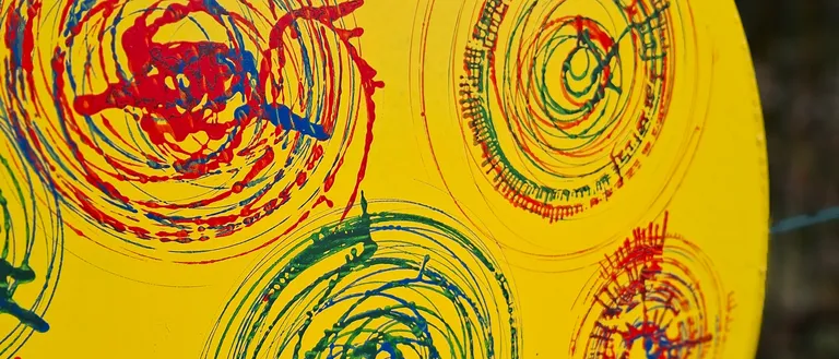 Ein gelbes rundes Objekt mit mehreren Spiralen in Rot, Blau und Grün bemalt. Die Spiralen sind unregelmäßig und haben unterschiedliche Dicke. Der Hintergrund ist gelb.