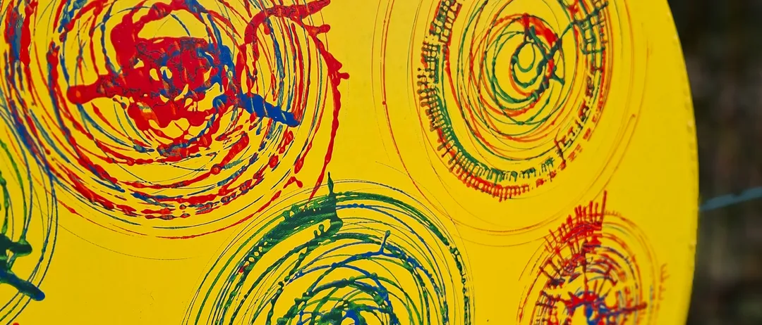 Ein gelbes rundes Objekt mit mehreren Spiralen in Rot, Blau und Grün bemalt. Die Spiralen sind unregelmäßig und haben unterschiedliche Dicke. Der Hintergrund ist gelb.