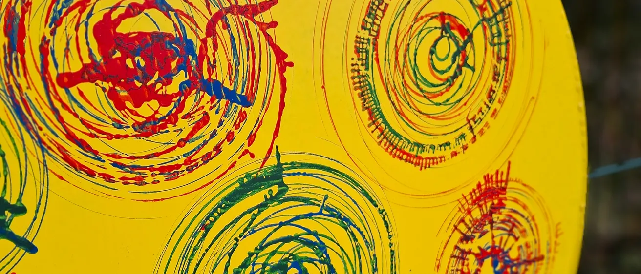 Ein gelbes rundes Objekt mit mehreren Spiralen in Rot, Blau und Grün bemalt. Die Spiralen sind unregelmäßig und haben unterschiedliche Dicke. Der Hintergrund ist gelb.