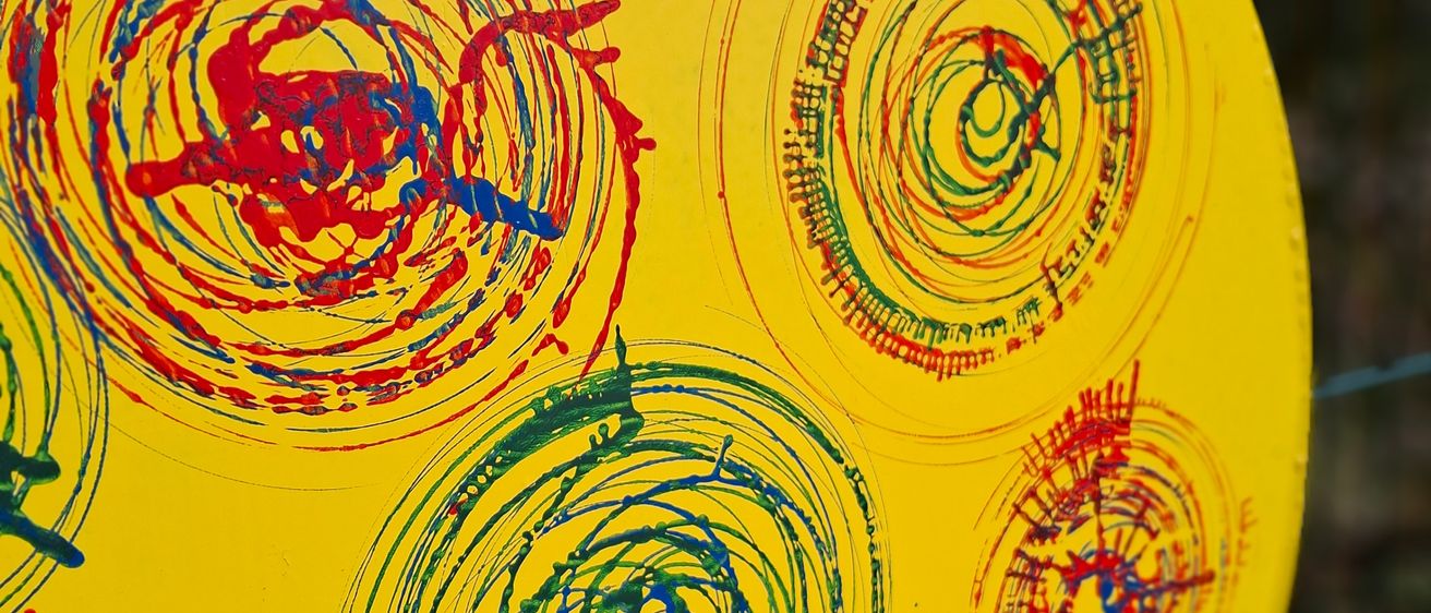 Ein gelbes rundes Objekt mit mehreren Spiralen in Rot, Blau und Grün bemalt. Die Spiralen sind unregelmäßig und haben unterschiedliche Dicke. Der Hintergrund ist gelb.