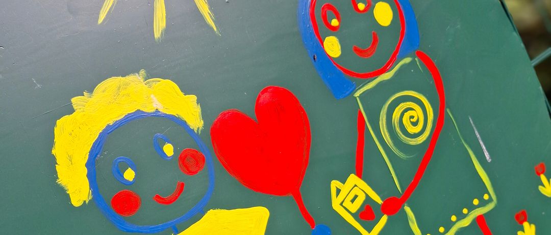 Ein grünes bemaltes Schild zeigt einen Jungen und ein Mädchen, die Händchen halten, mit einem Herzluftballon und Blumen um sie herum, unter einer Sonne.