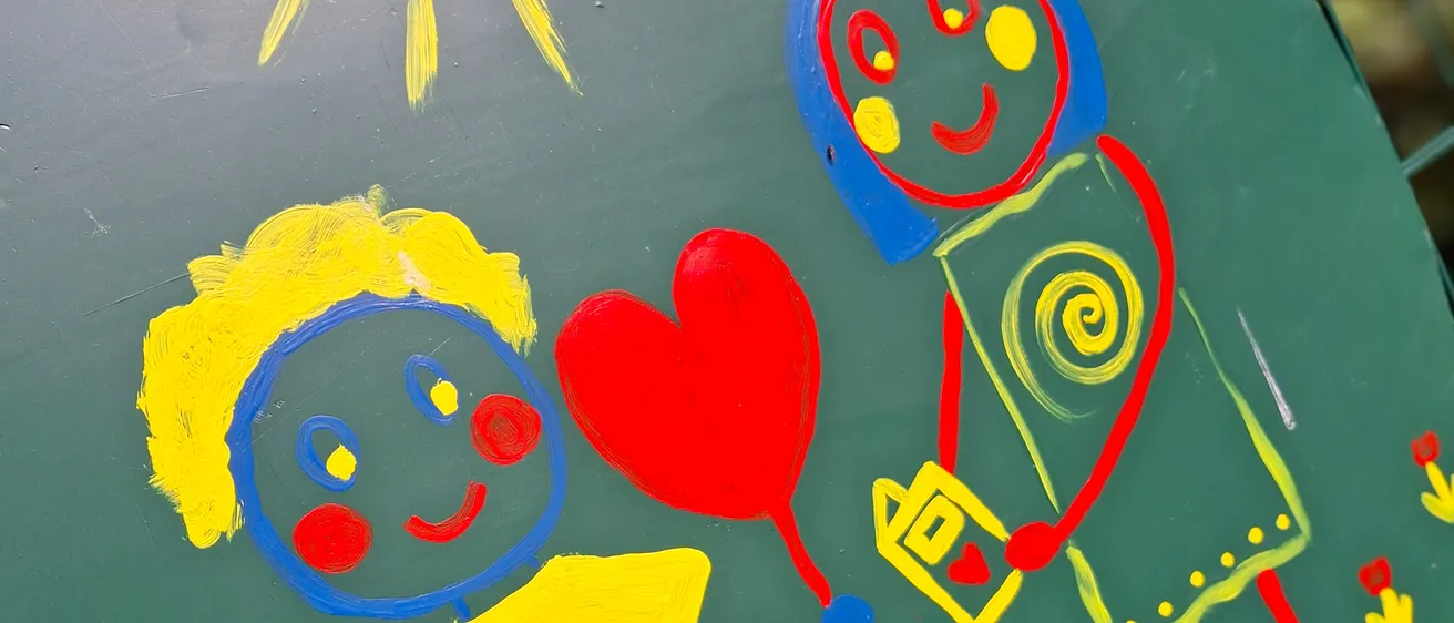 Ein grünes bemaltes Schild zeigt einen Jungen und ein Mädchen, die Händchen halten, mit einem Herzluftballon und Blumen um sie herum, unter einer Sonne.