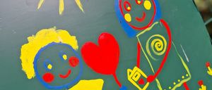 A green painted sign depicts a boy and a girl holding hands, with a heart balloon and flowers around them, under a sun.