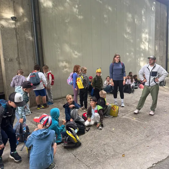 Eine Gruppe von Kindern, einige stehend und einige sitzend, versammelt sich vor einem Gebäude. Sie tragen Rucksäcke und Turnschuhe. Eine Frau und ein Mann stehen vor ihnen, und der Mann trägt einen Hut.
